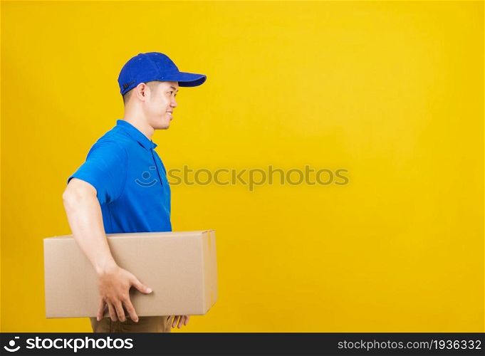 Portrait excited attractive delivery happy man logistic standing smile wearing blue t-shirt and cap uniform holding parcel box looking to camera, studio shot isolated on yellow background, side view