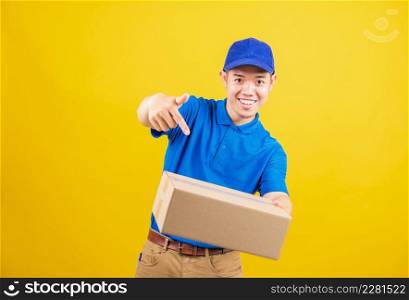 Portrait excited delivery happy man logistic standing he smile wearing blue t-shirt and cap uniform holding parcel box pointing finger to the box, studio shot isolated on yellow background