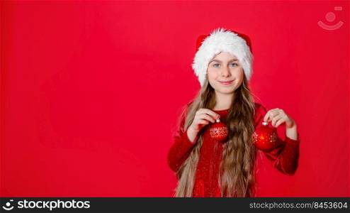 Portrait funny cheerful girl in a Santa hat and Christmas balls, isolated on a bright red background. The child points with his hand, place for the text.. Portrait funny cheerful girl in a Santa hat and Christmas balls, isolated on a bright red background.