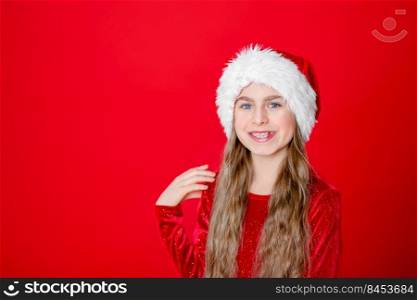 Portrait funny cheerful girl in a Santa hat and Christmas balls, isolated on a bright red background. The child points with his hand, place for the text.. Portrait funny cheerful girl in a Santa hat and Christmas balls, isolated on a bright red background.