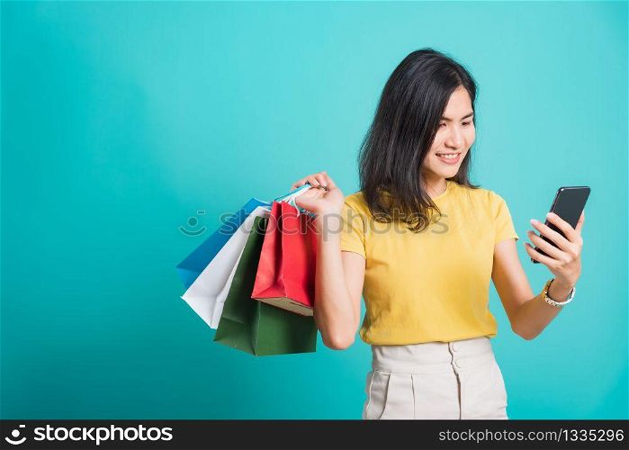 Portrait happy Asian beautiful young woman smile white teeth standing wear yellow t-shirt, She holding shopping bags and using a mobile phone, studio shot on blue background with copy space for text