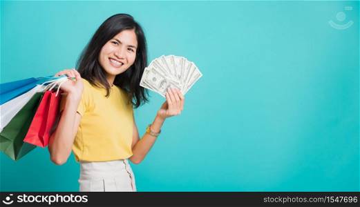 Portrait happy Asian beautiful young woman smile white teeth standing wear yellow t-shirt, She holding shopping bags and dollars money fan, studio shot on blue background with copy space for text