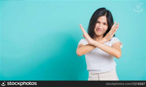 Portrait happy Asian beautiful young woman unhappy or confident standing wear white t-shirt, She holding two crossing arms say no X sign, studio shot on blue background with copy space for text