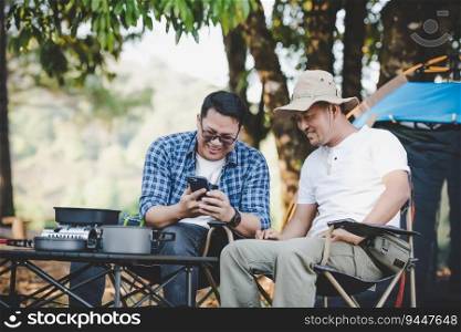 Portrait happy Asian man friends sitting on chair in the c&with talking and looking at smartphone. Cooking set front ground Outdoor cooking, traveling, c&ing, lifestyle concept.