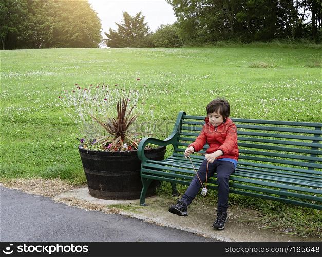 Portrait happy kid sitting alone on metal bench in the park, Child boy playing outdoor at green fields in spring or summer.