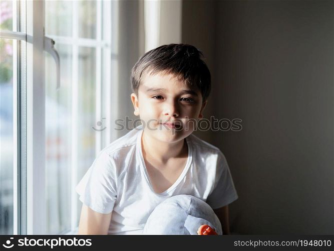 Portrait happy kid sitting next to window with autumn light in evening, High key light Cute young boy looking at camera with smiling face, Child boy sitting alone playing with toys at home on weekend