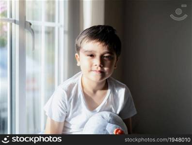 Portrait happy kid sitting next to window with autumn light in evening, High key light Cute young boy looking at camera with smiling face, Child boy sitting alone playing with toys at home on weekend