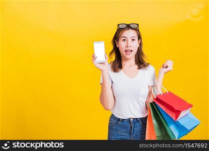 Portrait happy Thai Asian beautiful young woman smile standing, she surprises open mouth and holding shopping bags and show mobile phone blank screen, studio shot on yellow background with copy space