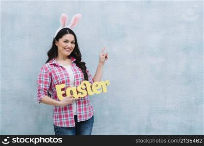 portrait happy woman with bunny ears head holding easter word pointing finger upward blue background