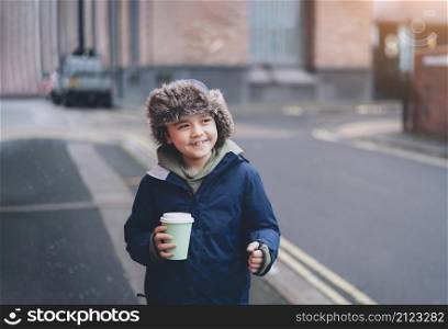 Portrait happy young boy looking out smiling face and holding cup of hot chocolate, Cute kid drinking hot drink while walking in the town,Child wearing a fur hooded winter coat having fun in the city in Autumn or Winter