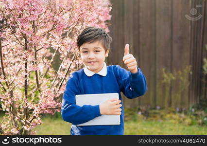 Portrait happy young boy showing thumbs up looking at camera with smiling face, School kid holding tablet pc standing outside waiting for School bus in sunny day Spring