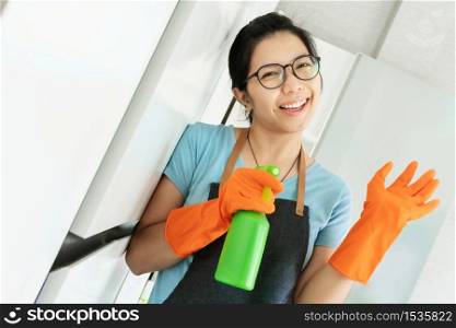 Portrait Happy Young girl in eyeglasses holding green cleaner spray bottle in white room. Smiling Asian Woman in Apron and orange rubber gloves preparing cleaning house. Having fun, Chores, Housework