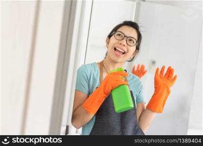Portrait Happy Young girl wear eyeglasses holding green cleaner spray bottle like at microphone singing in white room. Smiling Asian Woman in blue shirt with Apron and orange rubber gloves preparing cleaning her house. Having fun, Chores, Housework
