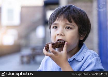 Portrait hugry little boy enjoy eating chocolate cake in outdoors cafe with blurry background of people, KId eating snack after playing at the park , Child eating food with yummy face