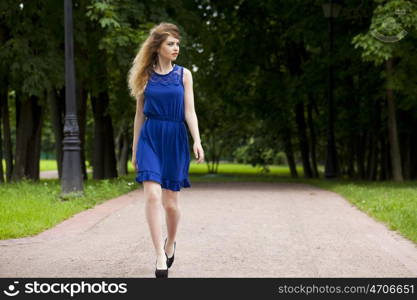 Portrait in full growth, attractive young blonde woman in blue dress walking in summer park