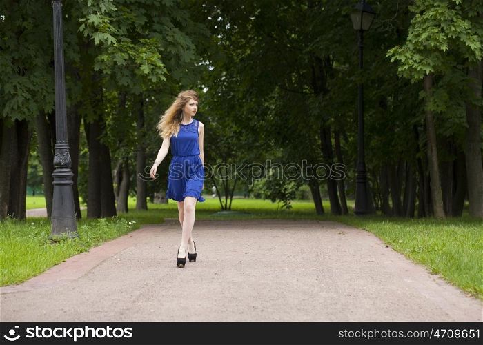 Portrait in full growth, attractive young blonde woman in blue dress walking in summer park