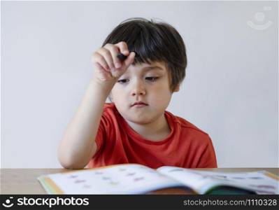 Portrait Kid boy holding black pen sitting alone and looking down with bored face ,Lonely child looking d down at table with sad face,Five years old kid bored with school homework,spoiled child