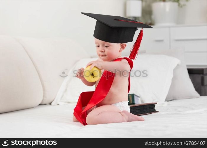 Portrait of 10 months old baby boy in graduation cap and ribbon holding yellow apple