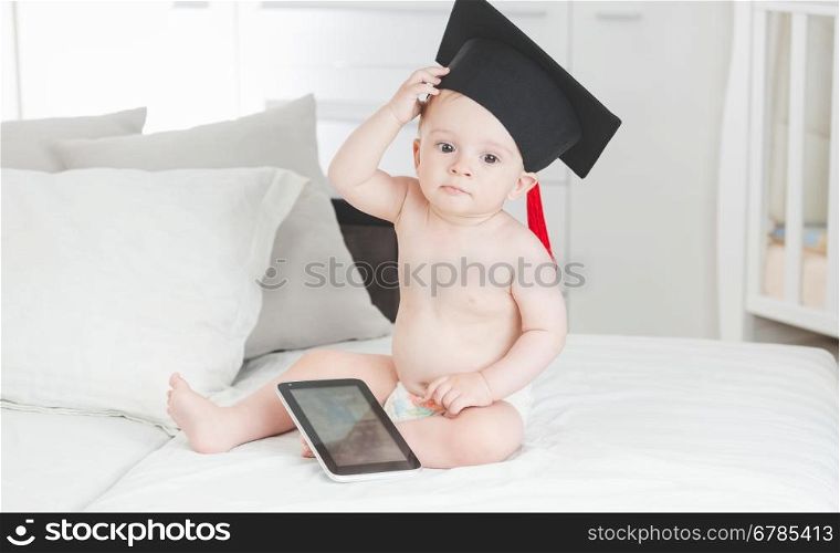 Portrait of 10 months old baby boy in graduation cap sitting with digital tablet on sofa at living room