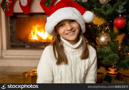 Portrait of 10 years old girl in Santa hat sitting next to fireplace and decorated Christmas tree