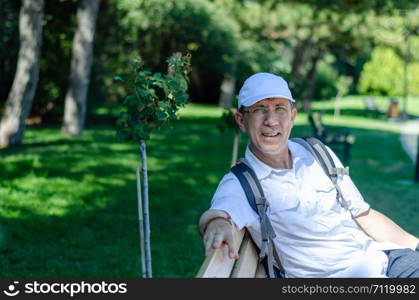 Portrait of 50s age man with glasses and sitting at the park.He has his backpack and hat.