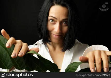 portrait of a beautiful adult woman with short black hair of a Caucasian appearance dressed in a white shirt holds a branch with green leaves, black background