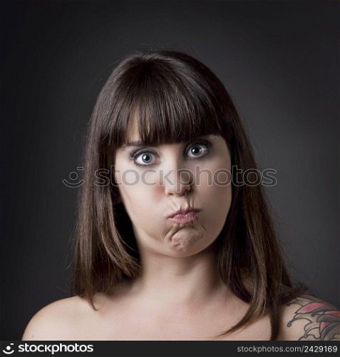 Portrait of a beautiful and funny woman with cheeks full of air against a grey background