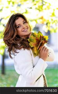 Portrait of a beautiful and lovely young woman smiling and holding fallen leaves on a blurred background of the autumn park on a sunny day.
