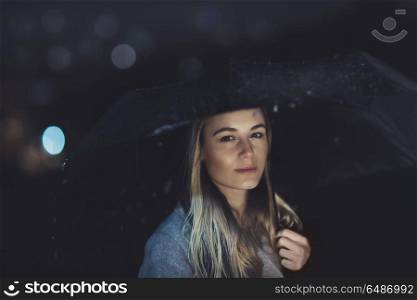 Portrait of a beautiful blond girl waiting for someone at dark rainy night, fashion women&rsquo;s portrait with umbrella over blurry night city lights background. Beautiful woman at rainy night