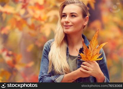 Portrait of a beautiful blond girl with dry tree leaves in hands dreamy looking far, standing over fall foliage background, enjoying autumn holidays in the park