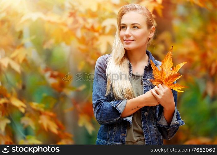 Portrait of a beautiful blond girl with dry tree leaves in hands dreamy looking far, standing over fall background, enjoying autumn holidays in the park