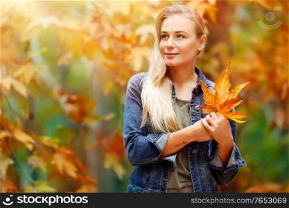 Portrait of a beautiful blond girl with dry tree leaves in hands dreamy looking far, standing over fall background, enjoying autumn holidays in the park