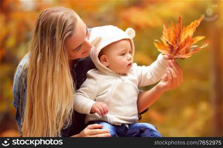 Portrait of a beautiful blond mother showing to her little son beauty of autumn nature, holding in hand dry orange maple leaves, spending day off in the fall park