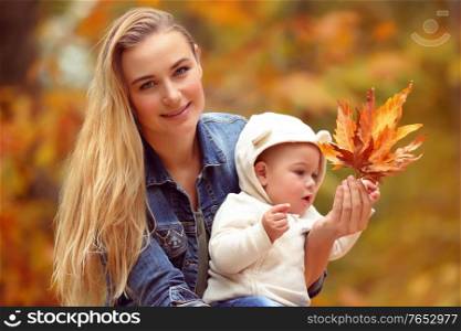 Portrait of a beautiful blond mother showing to her little son beauty of autumn nature, holding in hands dry orange maple leaves, spending time togather in the fall park