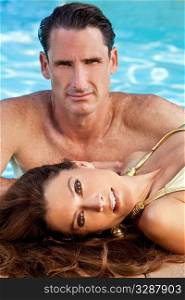 Portrait of a beautiful couple with a woman laying down at the side of a swimming pool with an attractive man behind her. The focus is on the woman in the foreground.