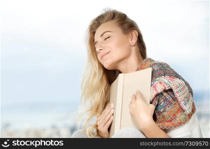 Portrait of a beautiful dreamy woman with closed eyes holding book near the heart, enjoying romantic story, happy quality time at home