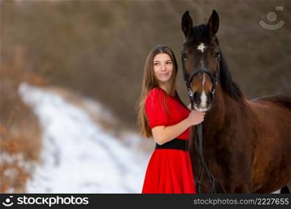 Portrait of a beautiful girl in a red dress standing with a horse on the background of a winter road