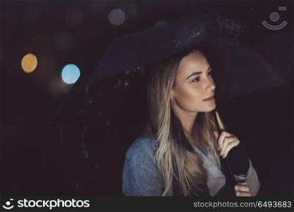 Portrait of a beautiful pensive woman standing alone with umbrella under rain in dark night and with sadness thinking about something. Pensive woman outdoors in rainy night