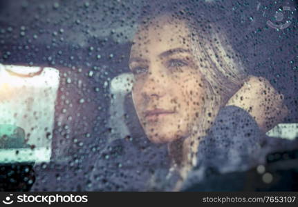 Portrait of a beautiful sad woman sitting in the car in rainy weather, pensive girl looking through the window glass with rain drops, autumn melancholy concept