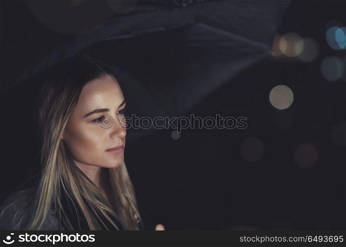 Portrait of a beautiful sad woman waiting for someone at night, standing with umbrella under rain over dark night background with bokeh city lights behind. Beautiful sad woman at rainy night
