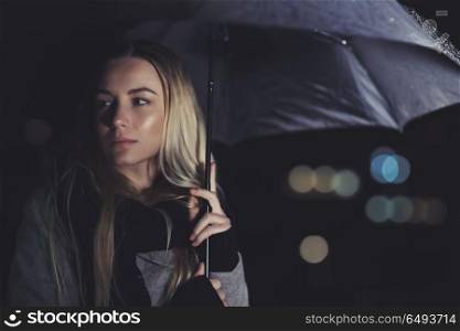 Portrait of a beautiful sad woman waiting for someone at night, standing with umbrella under rain over dark night background with bokeh lights behind. Beautiful sad woman at rainy night