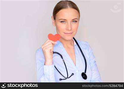 Portrait of a beautiful serious doctor wearing medical uniform and stethoscope isolated on gray background, holding in hands little red paper heart, healthy heart concept