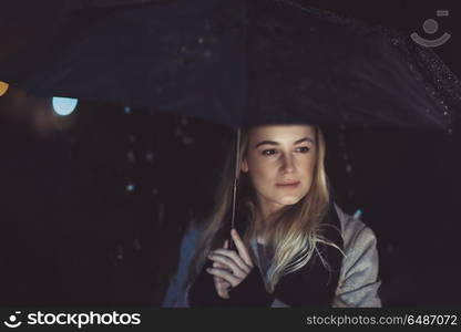 Portrait of a beautiful thoughtful woman standing alone with umbrella under the rain in dark night and with sadness thinking about something. Thoughtful woman outdoors on rainy night