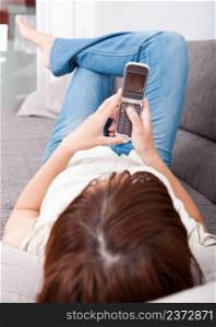 Portrait of a beautiful woman lying on sofa holding a cellphone and sending a sms