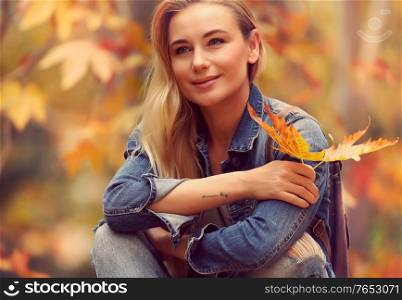 Portrait of a beautiful woman sitting in autumn park with dry maple leaves in hand, enjoying warm weather and beauty of fall nature