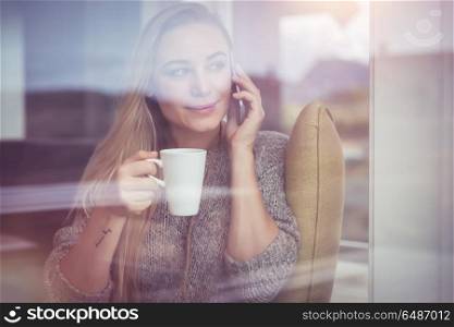 Portrait of a beautiful woman sitting in the country house with a cup of tea and speaking on the phone, looking through window, enjoying peaceful vacation. On the phone with cup of tea