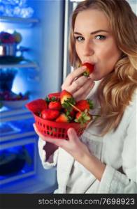 Portrait of a beautiful woman standing near the open refrigerator and eating a strawberry, with pleasure biting fresh sweet fruits, healthy organic nutrition