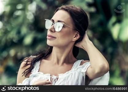 Portrait of a beautiful woman wearing stylish sunglasses over exotic plants background, luxury photoshoot on the tropical beach. Beautiful woman portrait