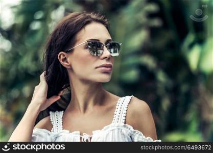 Portrait of a beautiful woman wearing stylish sunglasses over exotic plants background, luxury photoshoot on the tropical beach, fresh tropical breeze . Tropical breeze