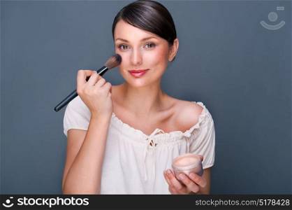 Portrait of a Beautiful woman with makeup brush near her face looking at camera and smiling against grey background